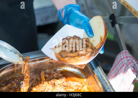 white bread bun in a white food tray being topped with toppings such as pulled pork and coleslaw with gloved hands holding tongs Stock Photo