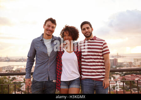 Three happy young friends posing for a picture on a bridge with a city behind them and very bright white clouds Stock Photo