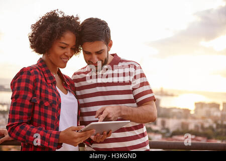 Young couple looking at tablet while he points at something on it while the sun is setting behind them over a city scape Stock Photo