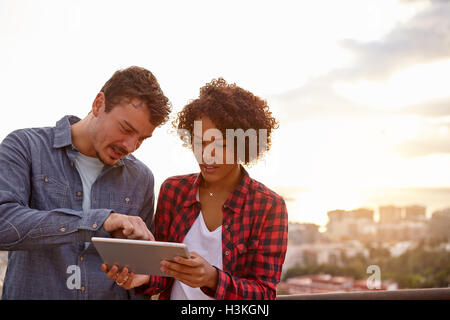 Young couple looking at a tablet the girl is holding and the guy is pointing at with the sun setting behind them Stock Photo