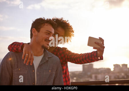 Young couple taking a selfie with her arm around his neck and a big toothy smile while looking at the cellphone Stock Photo