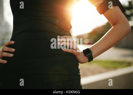 Cropped shot of fit woman in sports wear standing with her hand on hip outdoors, with sun flare. Sportswoman wearing smartwatch Stock Photo