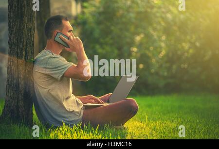 Outdoor Office Worker. Men Making Business Calls While Seating on the Grass in a Park with Laptop Computer on His Legs. Out of O Stock Photo