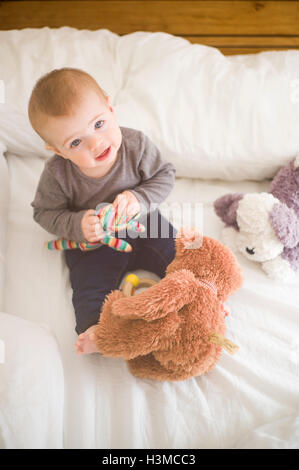 Baby girl sitting on bed holding soft toys looking up at camera Stock Photo