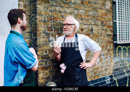 Senior craftsman drinking coffee and chatting to young man outside workshop Stock Photo