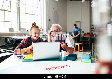 Senior male craftsman laughing and looking at laptop with young woman in book arts workshop Stock Photo
