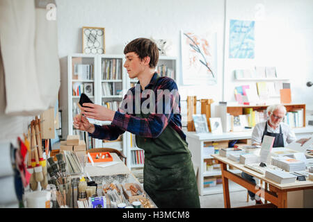 Young craftsman picking up brush and looking at materials in craft shop, with senior man in background Stock Photo