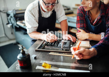 Senior craftsman using letterpress equipment and teaching young craftsman and craftswoman in print workshop Stock Photo