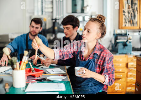 Young craftswoman holding coffee cup and reaching out for paintbrush in creative print studio, with two craftsmen in background Stock Photo