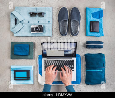Overhead view of man's hands typing on laptop surrounded by travel packing items, with blue shirt,  retro camera, wash bag and notebook Stock Photo