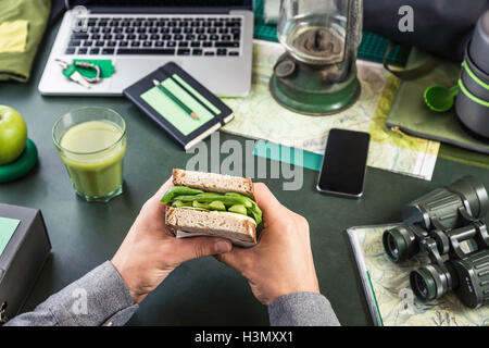 Mans hand's holding sandwich at table with hiking equipment and laptop Stock Photo