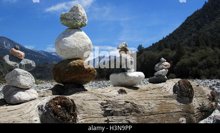 Stone pile cairns in riverbed balancing on a driftwood log with mountains in the background Stock Photo