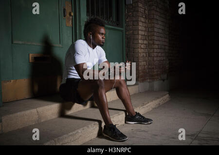 Young man with mohawk, sitting on steps at night, wearing earphones Stock Photo
