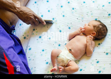 Newborn baby boy lying on bed next to father using smartphone Stock Photo