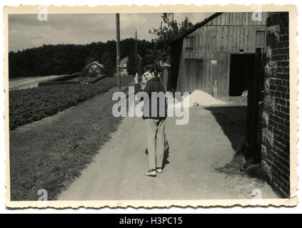 BERLIN, GERMANY - CIRCA 1963: Woman walking along the path past the brick building of a wooden barn, half turning greets photographer who is behind her Stock Photo