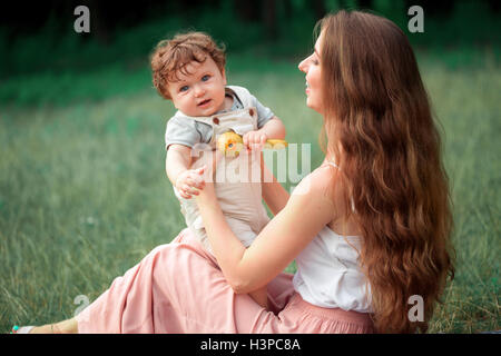 Young beautiful mother sitting with her little son against green grass. Happy woman with her baby boy on a summer sunny day. Family walking on the meadow. Stock Photo