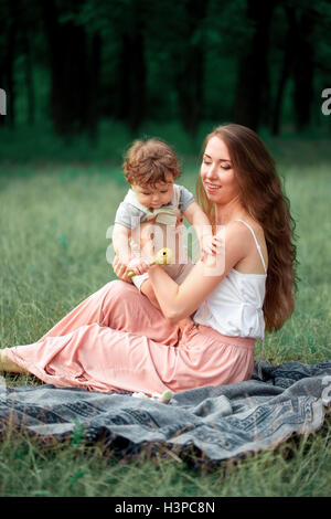 Young beautiful mother sitting with her little son against green grass. Happy woman with her baby boy on a summer sunny day. Family walking on the meadow. Stock Photo