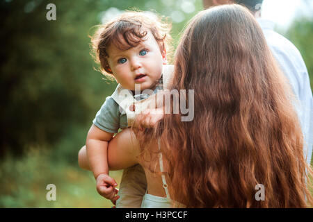 Young beautiful mother hugging her little toddler son against green grass. Happy woman with her baby boy on a summer sunny day. Family walking on the meadow. Stock Photo