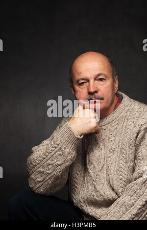 elderly man sits and looks thoughtfully, pondering problem or complex issue in sweater. Stock Photo