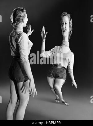 1940s 1950s YOUNG BLOND LAUGHING WOMAN LOOKING AT HERSELF DISTORTED IN FUN HOUSE MIRROR Stock Photo