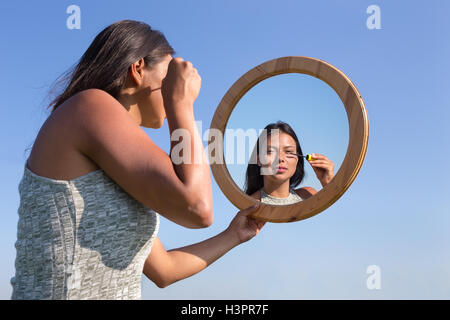 Woman applying cosmetics mascara makeup in mirror outside with blue sky Stock Photo