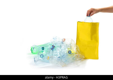 Pile of plastic bottles is lying on the white background. Woman's hand is inserting plastic bottle into an empty yellow bag. Stock Photo