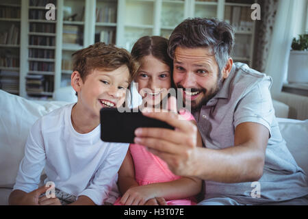 Father sitting on sofa with his children and clicking a selfie in living room Stock Photo