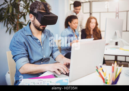 Businessman wearing virtual reality headset while using laptop against colleagues Stock Photo