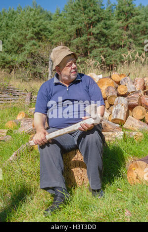 Senior man sitting on a log getting ready to chop pile of firewoods for winter kindling Stock Photo