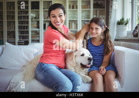 Mother sitting on sofa and tying daughters hair in living room Stock Photo
