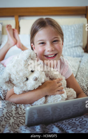 Happy girl lying on bed with a teddy bear and using digital tablet Stock Photo