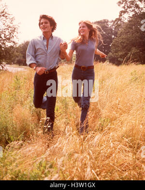 1970s SMILING HAPPY COUPLE RUNNING THROUGH MEADOW GRASS WEARING BLUE SHIRTS DENIM JEANS Stock Photo