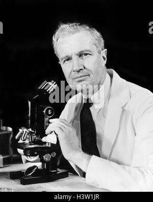 1940s MAN SCIENTIST USING MICROSCOPE WEARING A WHITE LAB COAT AND TIE LOOKING AT CAMERA Stock Photo