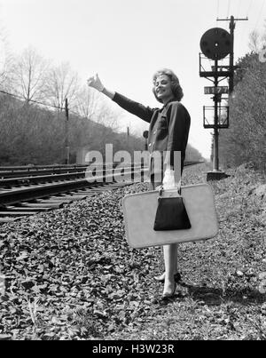 1950s 1960s WOMAN SMILING HOLDING SUITCASE STANDING BESIDE RAILROAD TRACKS GLOVED HAND THUMB IN THE AIR TO HITCH A RIDE Stock Photo