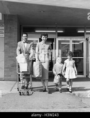 1950s FAMILY OF FOUR WALKING OUT OF GROCERY STORE FATHER PUSHING SHOPPING CART MOTHER AND SON CARRYING BAGS Stock Photo