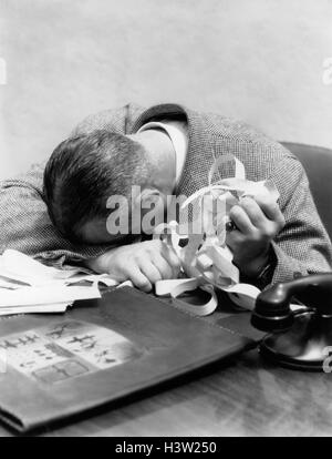 1930s 1940s MAN WITH HEAD DOWN ON DESK AND STOCK TICKER TAPE IN HAND Stock Photo