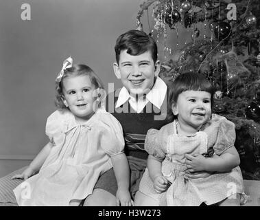 1940s 1950s SMILING BIG BROTHER AND TWO SISTERS SITTING TOGETHER BY CHRISTMAS TREE Stock Photo