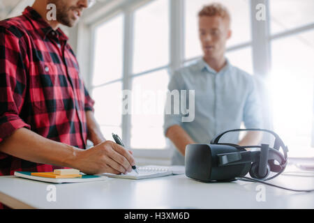 Shot of two young man standing at a table with VR goggles writing on notepad. Developers testing virtual reality glasses in offi Stock Photo