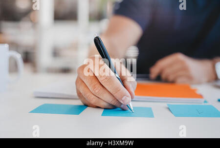 Close up image of business man writing on an adhesive note at table in office, focus on hand and pen. Stock Photo