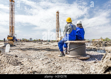 Two construction workers with helmets are sitting on wooden spool with steel cable Stock Photo