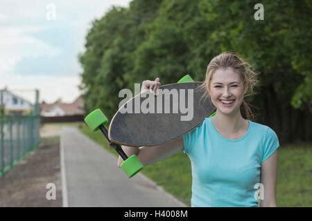 Portrait of cheerful woman holding skateboard standing on footpath at park Stock Photo