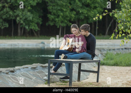 Loving man resting with girlfriend playing guitar on park bench by lake Stock Photo