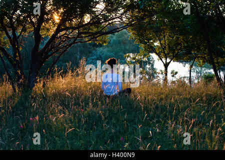 Rear view of woman meditating on grassy field by trees Stock Photo