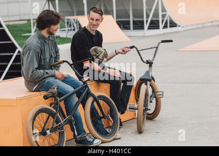 Cheerful friends resting on seat with bicycles at skateboard park Stock Photo