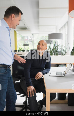 Smiling confident businessmen listening to male colleague at desk in creative office Stock Photo