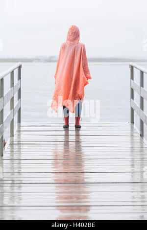 Rear view of woman wearing raincoat standing on jetty during rainy season Stock Photo