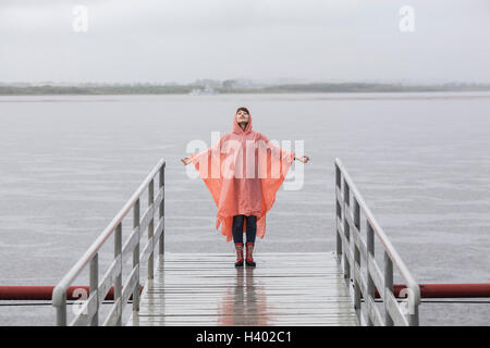 Woman wearing raincoat enjoying rainy season while standing on jetty Stock Photo