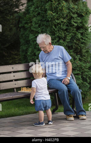 Grandfather playing chess with girl on park bench against tree Stock Photo