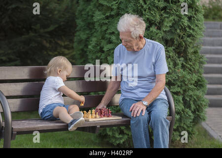 Senior man playing chess with granddaughter on park bench against tree Stock Photo
