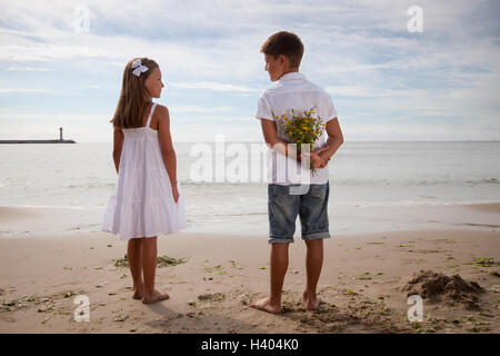 Boy holding flowers behind his back, standing next to girl on beach